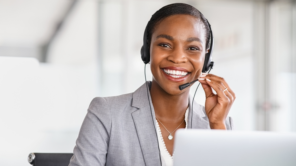 A professional female agent wearing a headset and smiling while providing Cold Calling Services on a laptop in a modern office setting.